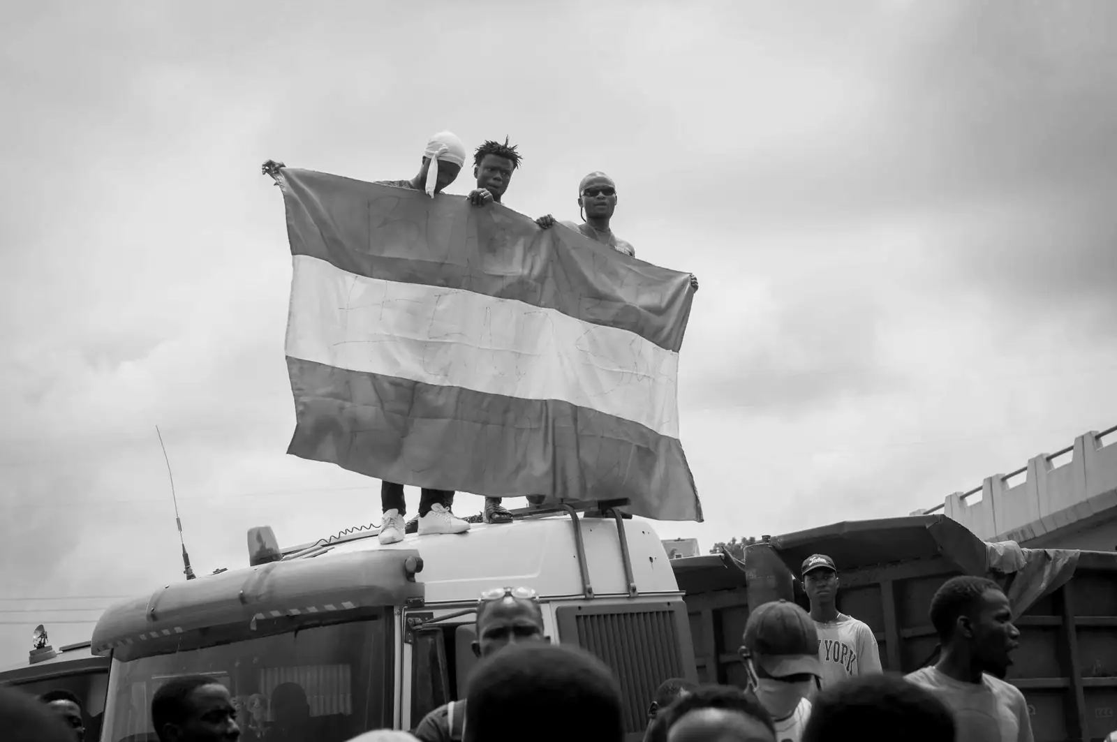 Three men on top of a truck hold a large flag up above a crowd.
