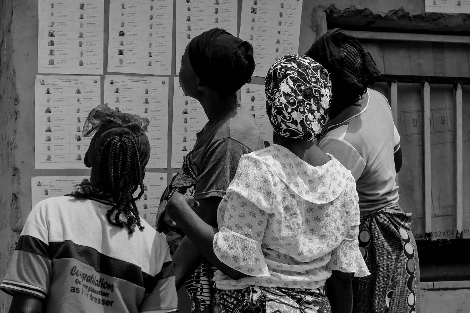 Four women, seen from behind, read printed voter lists pasted on a wall.
